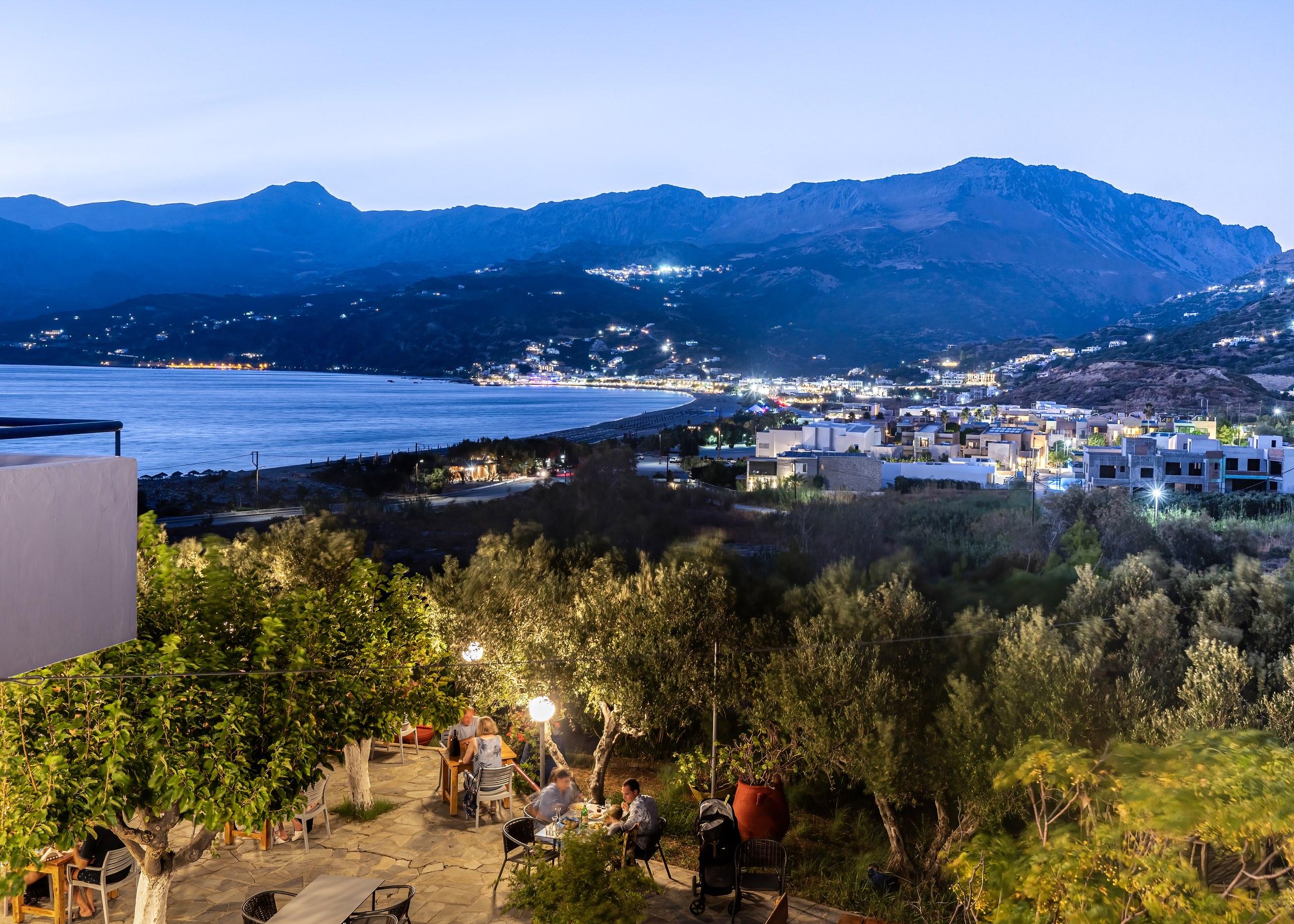 The garden terrace at dusk, tables lit under the olive trees with the bay behind